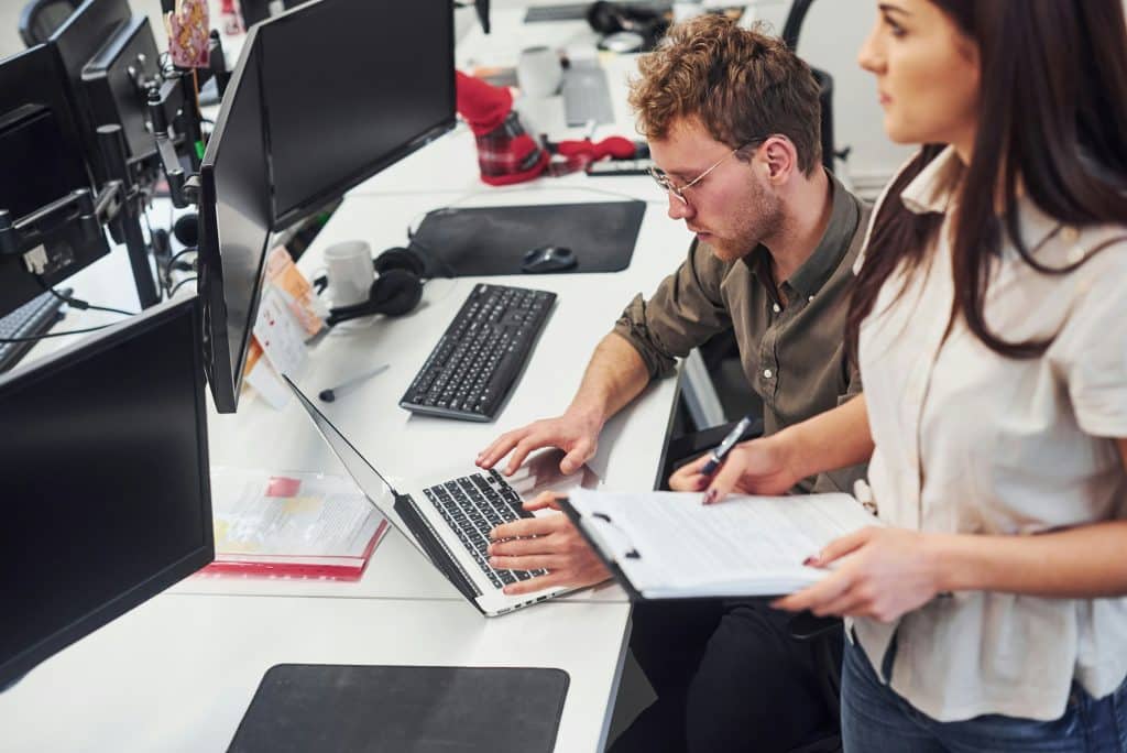 Two researchers collaborating at an office desk, analyzing data on a laptop and a clipboard.