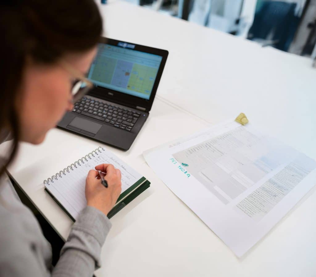 A person takes handwritten notes in a notebook while looking at an open laptop on a desk.